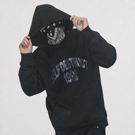 A person wearing a black hoodie with 'SELF DESTRUCT 1993' printed in gray on the front, holding up the hood to reveal a black and white bandana over the face, against a plain light gray background.