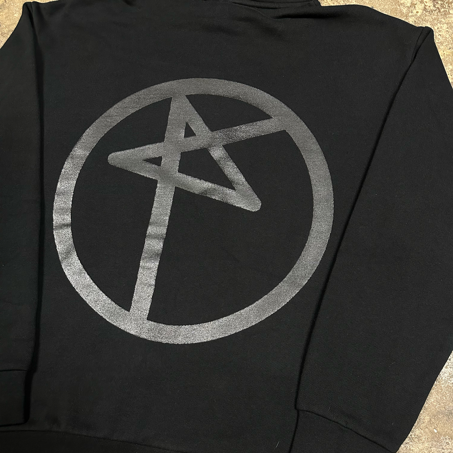 Black hoodie with a silver symbol on the back, laid flat on a textured surface.