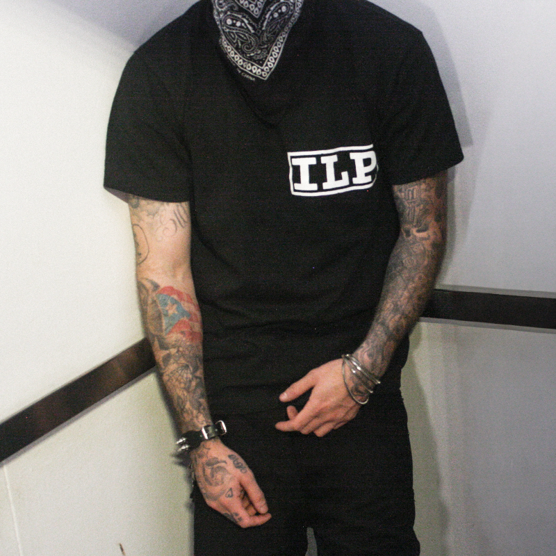 A tattooed man wearing a black short-sleeve t-shirt with a white 'ILP' logo on the chest, paired with black pants and a black-and-white bandana covering his face, standing against a white wall.