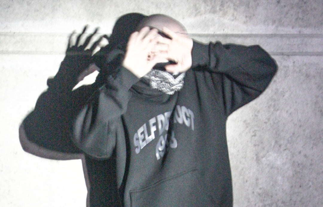 A person wearing a dark gray hoodie with 'SELF DESTRUCTION 1998' printed on the front, standing against a textured gray wall with their hands covering their face and casting a long shadow.