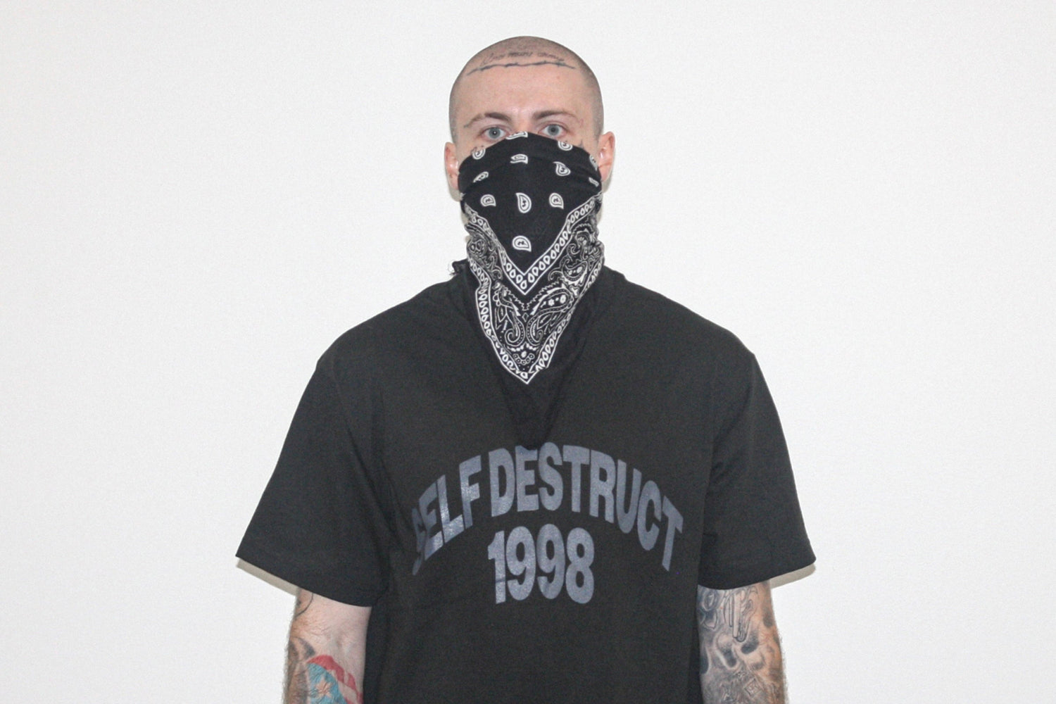 A man wearing a black bandana with a white paisley pattern over his mouth and nose, paired with a black t-shirt that reads 'SELF DESTRUCT 1998' in gray lettering, set against a plain white background.
