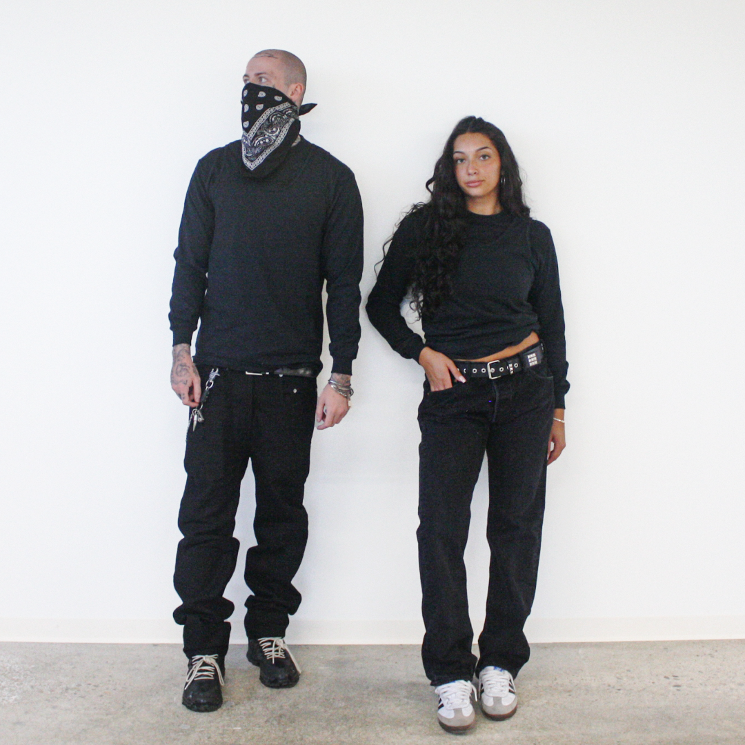 A man and a woman standing side-by-side against a white wall, both wearing black long-sleeve crewneck shirts, black jeans, and black belts. The man also wears a black and white patterned bandana covering his mouth and neck, while the woman wears white sneakers.