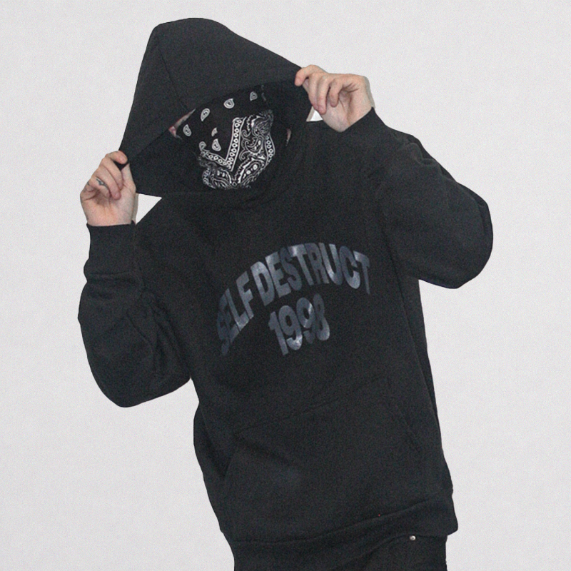 A person wearing a black hoodie with 'SELF DESTRUCT 1993' printed in gray on the front, holding up the hood to reveal a black and white bandana over the face, against a plain light gray background.