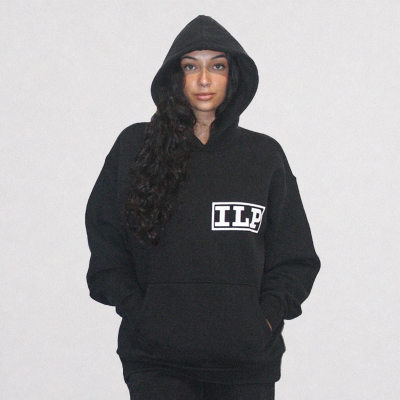 A woman wearing a black pullover hoodie with a white outlined 'ILP' logo on the chest, posed against a light gray studio background.