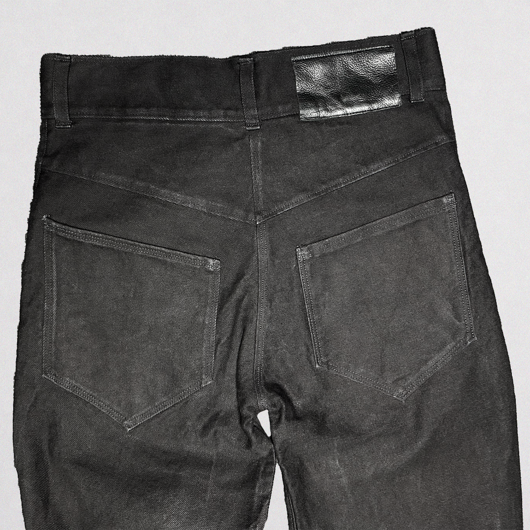 Rear view of dark gray washed denim jeans showing the back pockets, belt loops, and a black leather patch on the waistband, set against a light gray background.