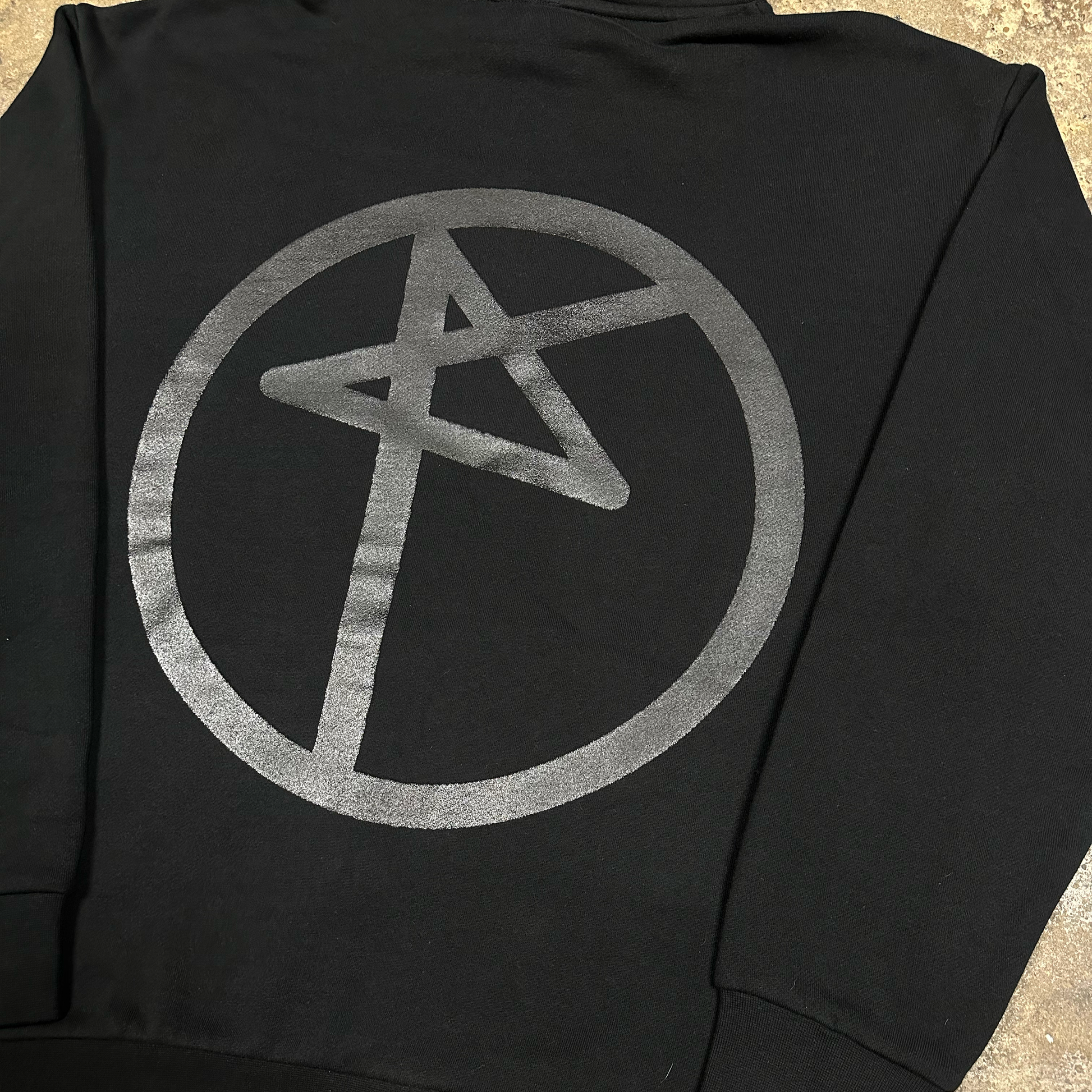 Black hoodie with a silver symbol on the back, laid flat on a textured surface.