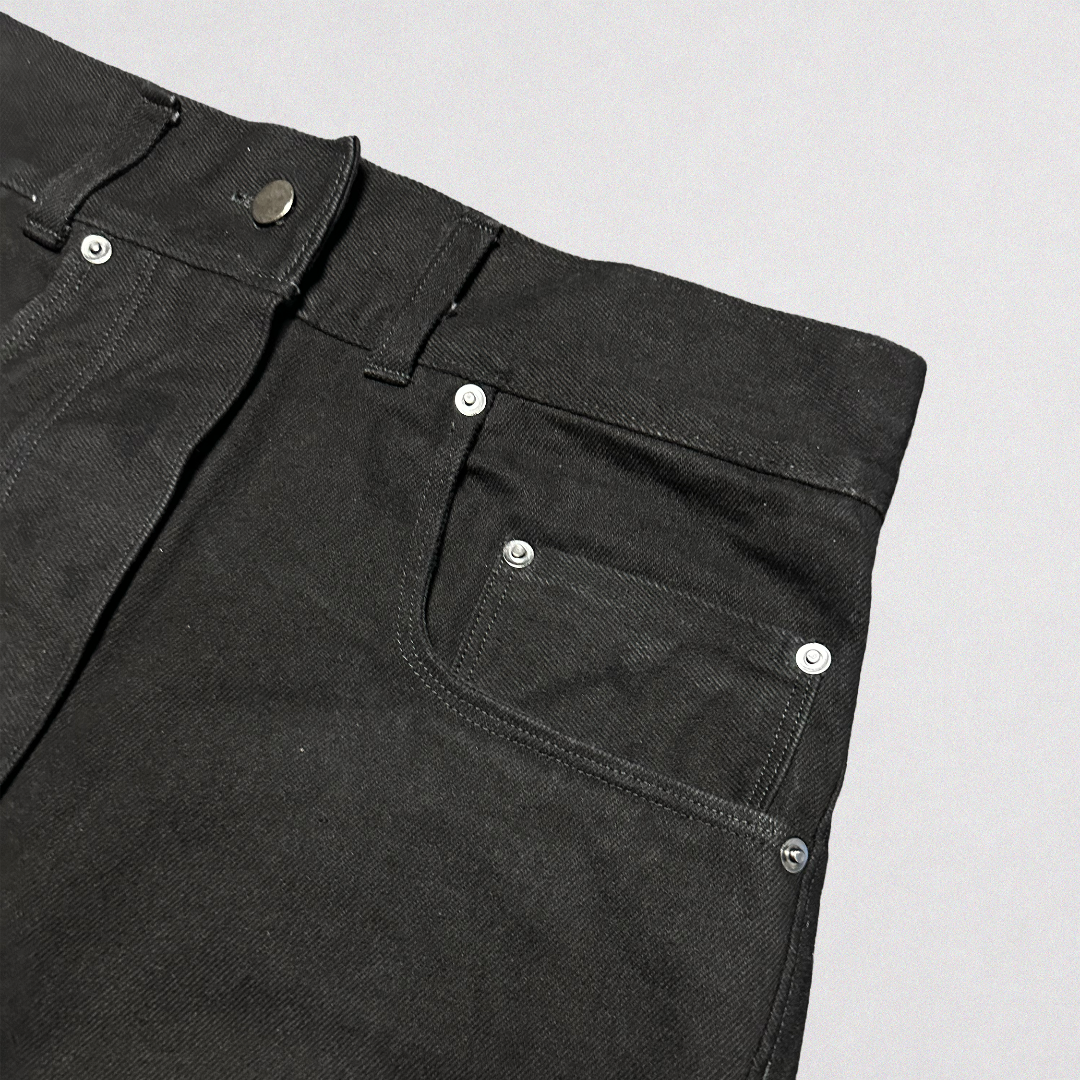 Close-up of black denim jeans showing the waistband, belt loops, front pocket, and silver rivets on a light gray background.