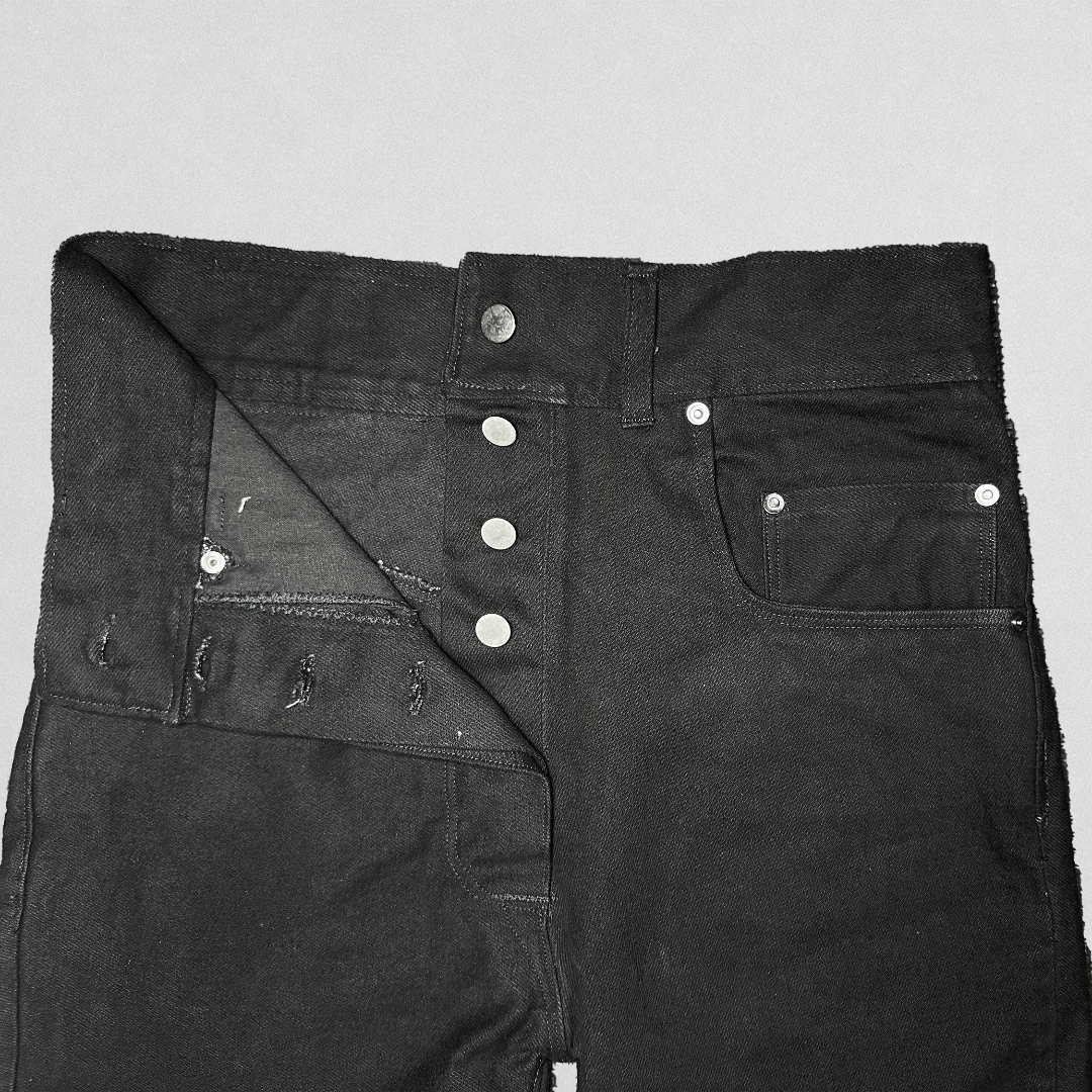 Close-up of a pair of black denim jeans laid flat on a light gray background, showing the front waistband, button closure, pocket details, and raw hem.