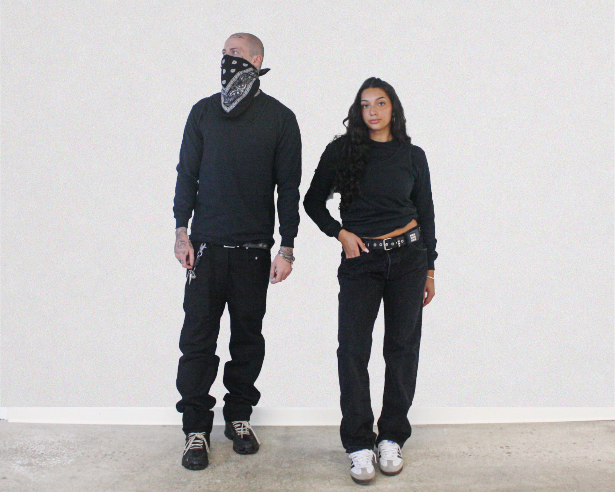 Two models standing against a plain white wall, both wearing black long-sleeve shirts, black jeans, and black belts. The model on the left also wears a black bandana over his face and black sneakers.