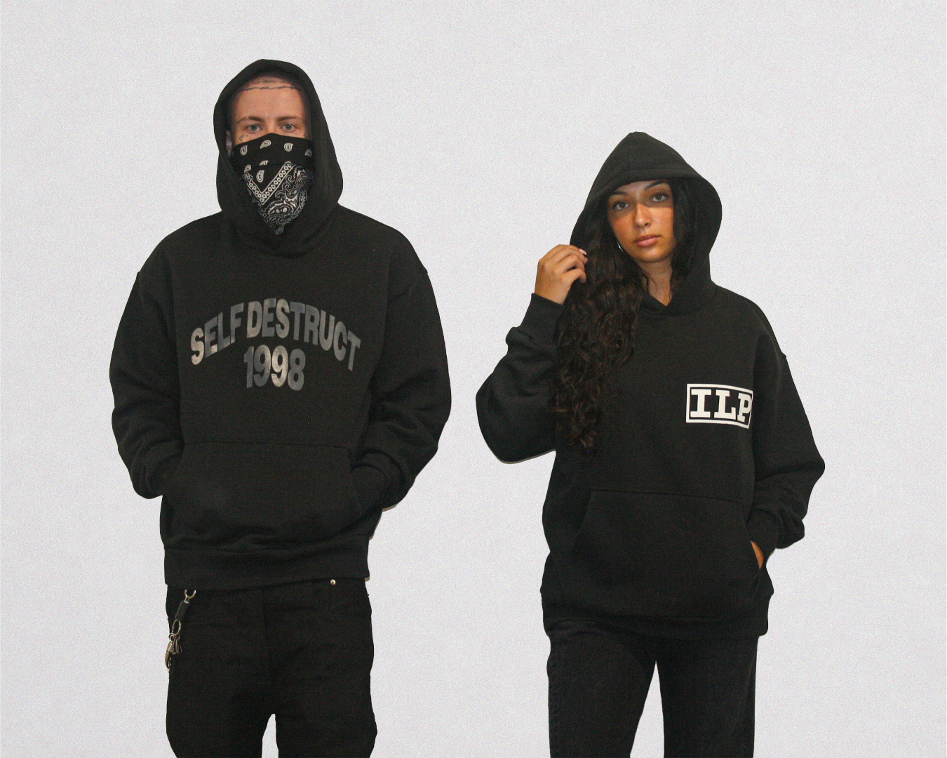 Two models wearing black hoodies against a light gray background. The model on the left wears a black bandana over their face and a black hoodie with 'SELF DESTRUCT 1998' printed in gray on the chest.