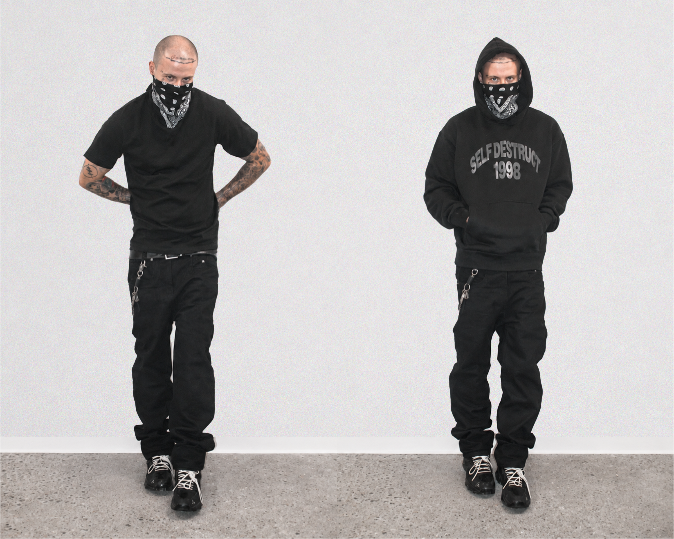 Two men modeling black streetwear outfits against a plain light gray wall and concrete floor. The left model wears a black t-shirt, black pants, and a black bandana.