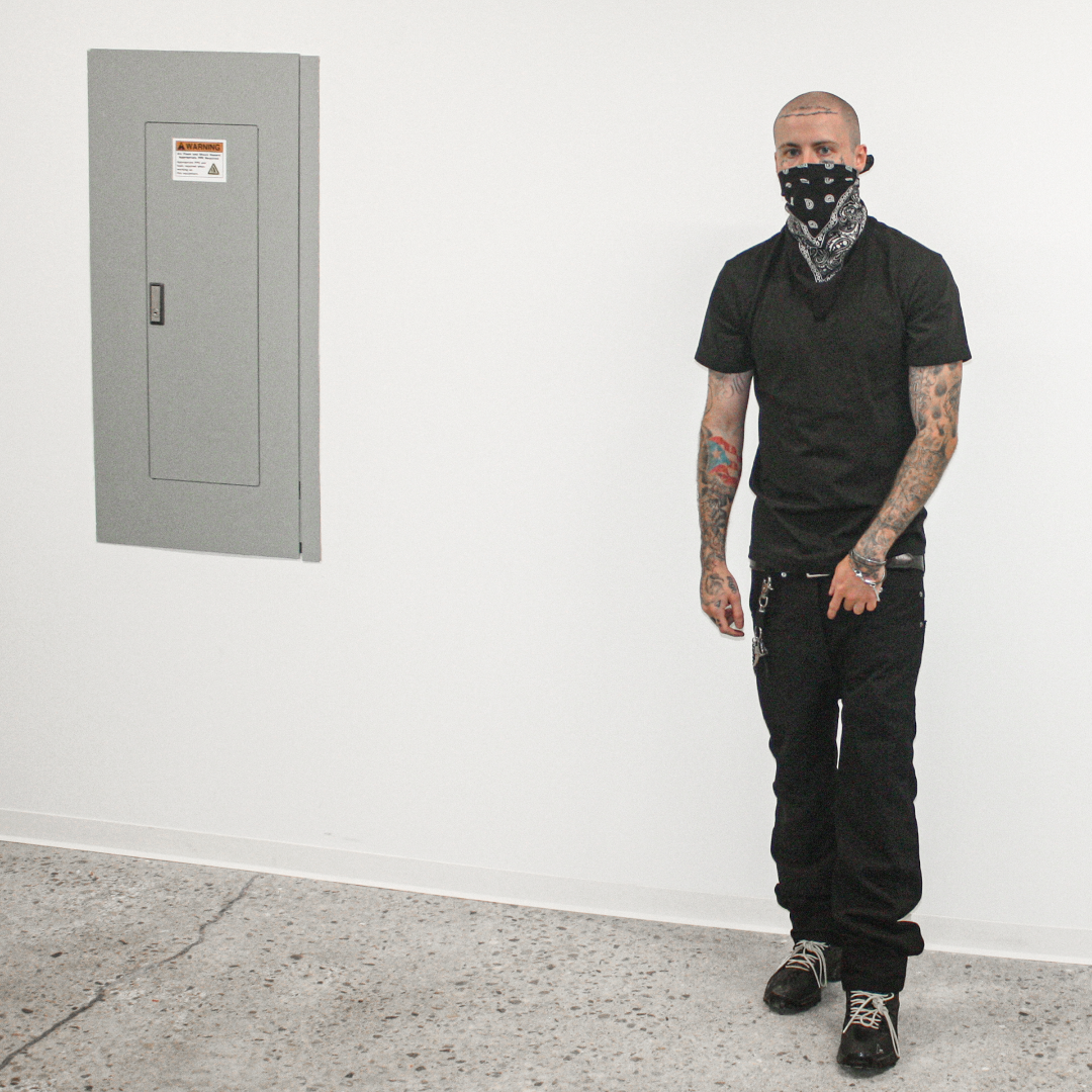 A tattooed man wearing a black t-shirt, black cargo pants, and a black-and-white bandana stands in a minimalist white room with a gray electrical panel on the wall.