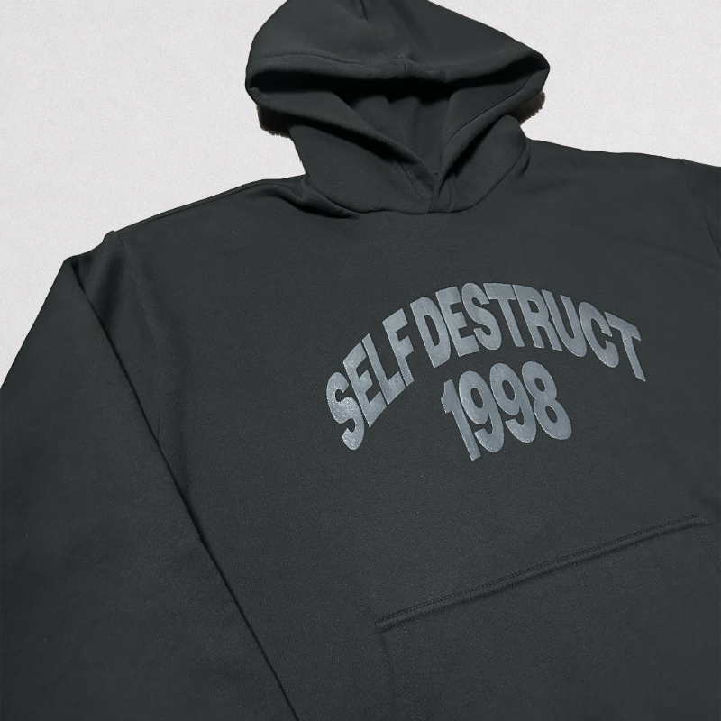 A dark charcoal gray hoodie featuring the words 'SELF DESTRUCT 1998' printed in a light gray arched font on the chest, shown against a neutral light gray background.