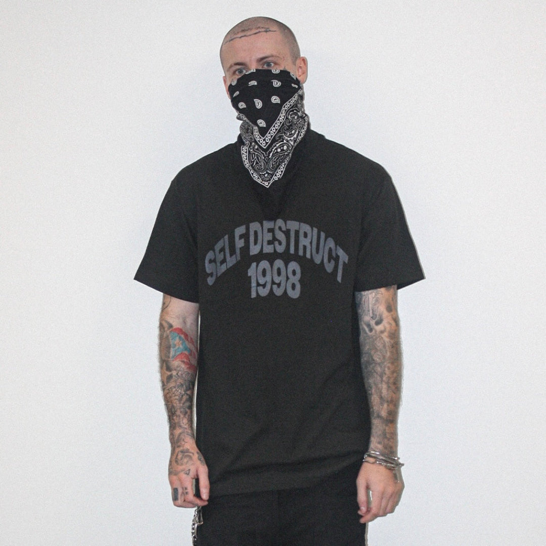 A man with tattoos and a bandana over his mouth wears a black t-shirt with 'SELF DESTRUCT 1998' printed in gray on the front, set against a plain white background.