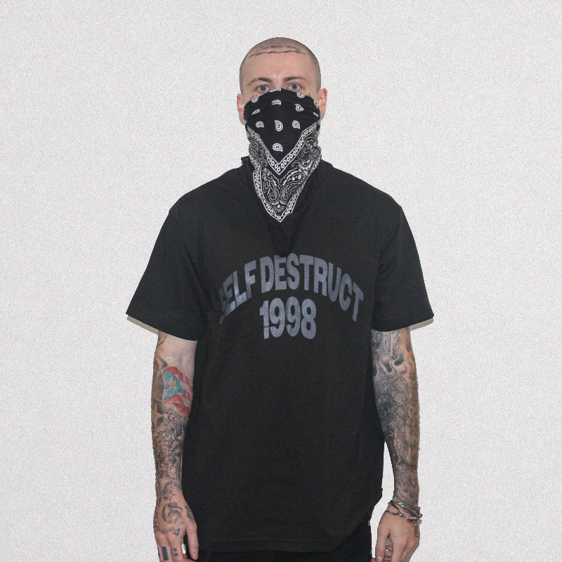 A tattooed man wearing a black bandana over his nose and mouth, and a black graphic t-shirt with 'SELF DESTRUCT 1998' printed in gray on the front, standing against a plain light gray background.